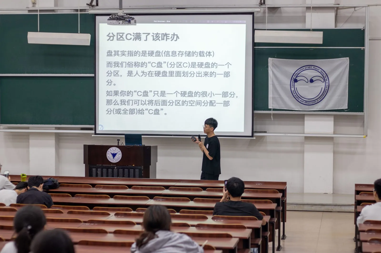 讲座投影上写着“分区C满了该咋办”，正解释“盘”指硬盘、而 C 盘只是硬盘上人为划出的一个分区；讲台右侧挂着协会队旗
