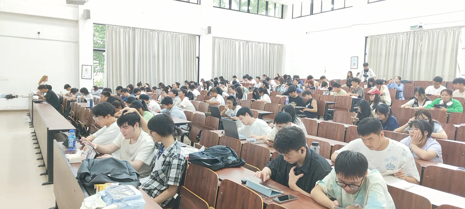 从讲台方向拍的阶梯教室：长条木桌一排排向后延伸，几乎每个座位都坐了人，多数低头看着自己的笔记本电脑或手机；前方两侧墙上各挂一块投影幕，窗外是白天的天光