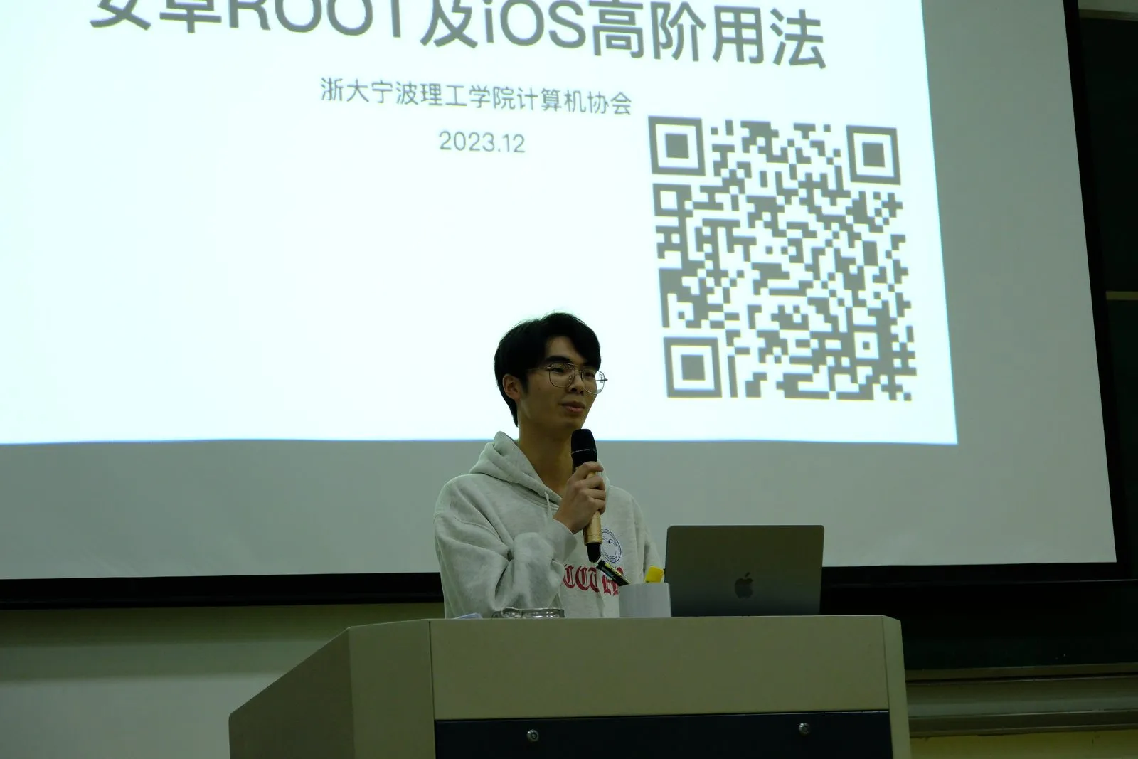 讲台上一名成员持话筒讲解，身后投影的标题页写着“安卓 ROOT 及 iOS 高阶用法／浙大宁波理工学院计算机协会／2023.12”，右侧有一个二维码