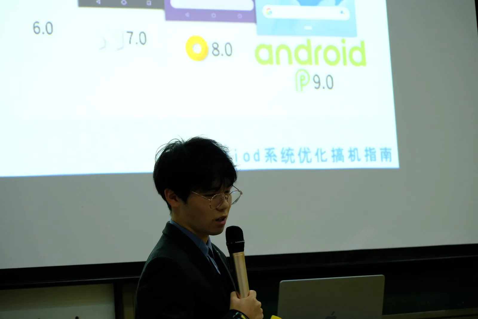 投影上并排展示 Android 6.0、7.0、8.0 与 Android P 9.0 的图标与界面，下方标题为系统优化搞机指南，讲者在画面前侧身讲解