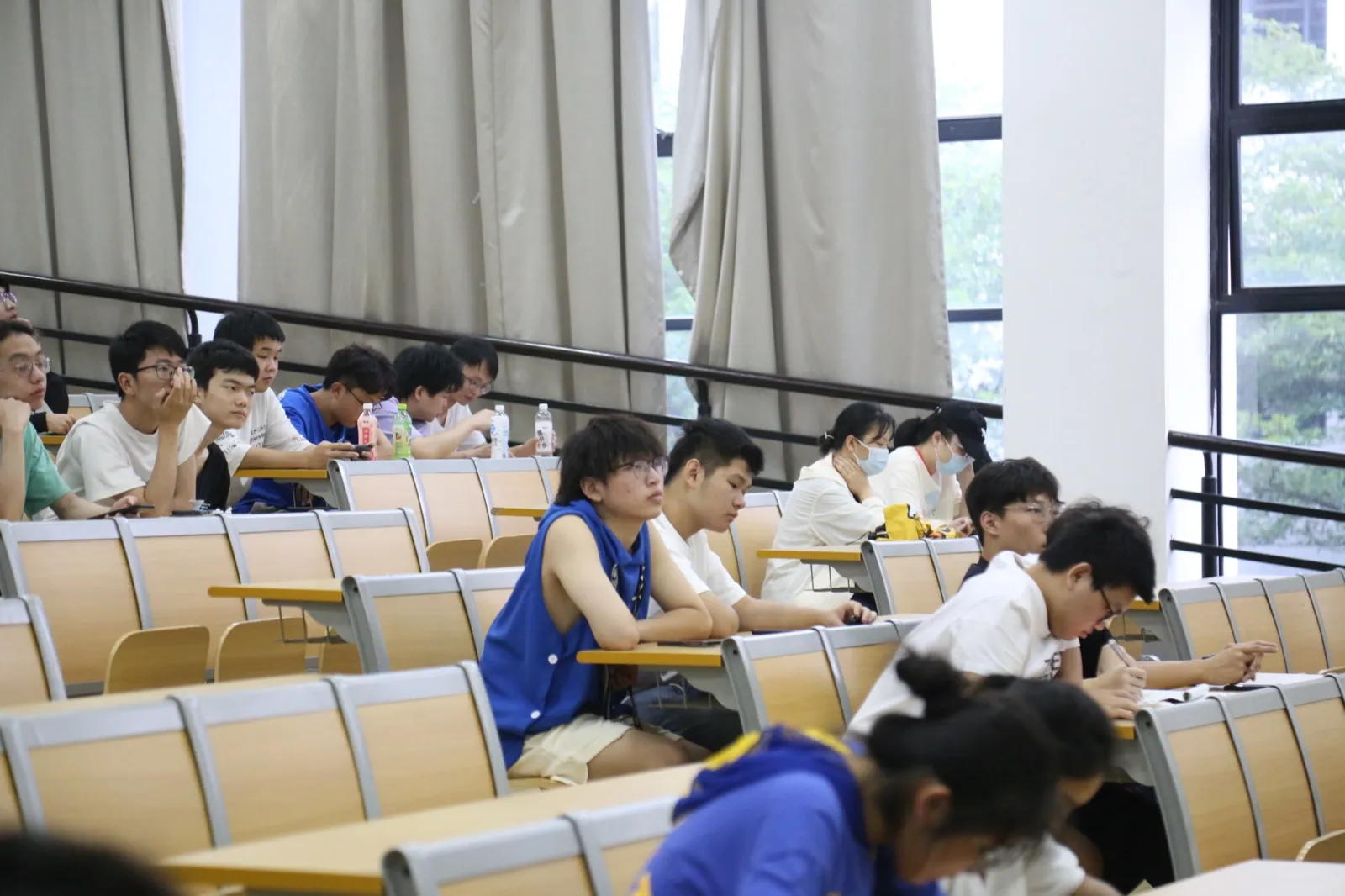 阶梯教室里学生分散坐在几排座位上,多数侧身朝向讲台方向,有人托腮听讲,有人低头做笔记,桌上放着饮料瓶;右侧是整面落地窗,窗外绿树