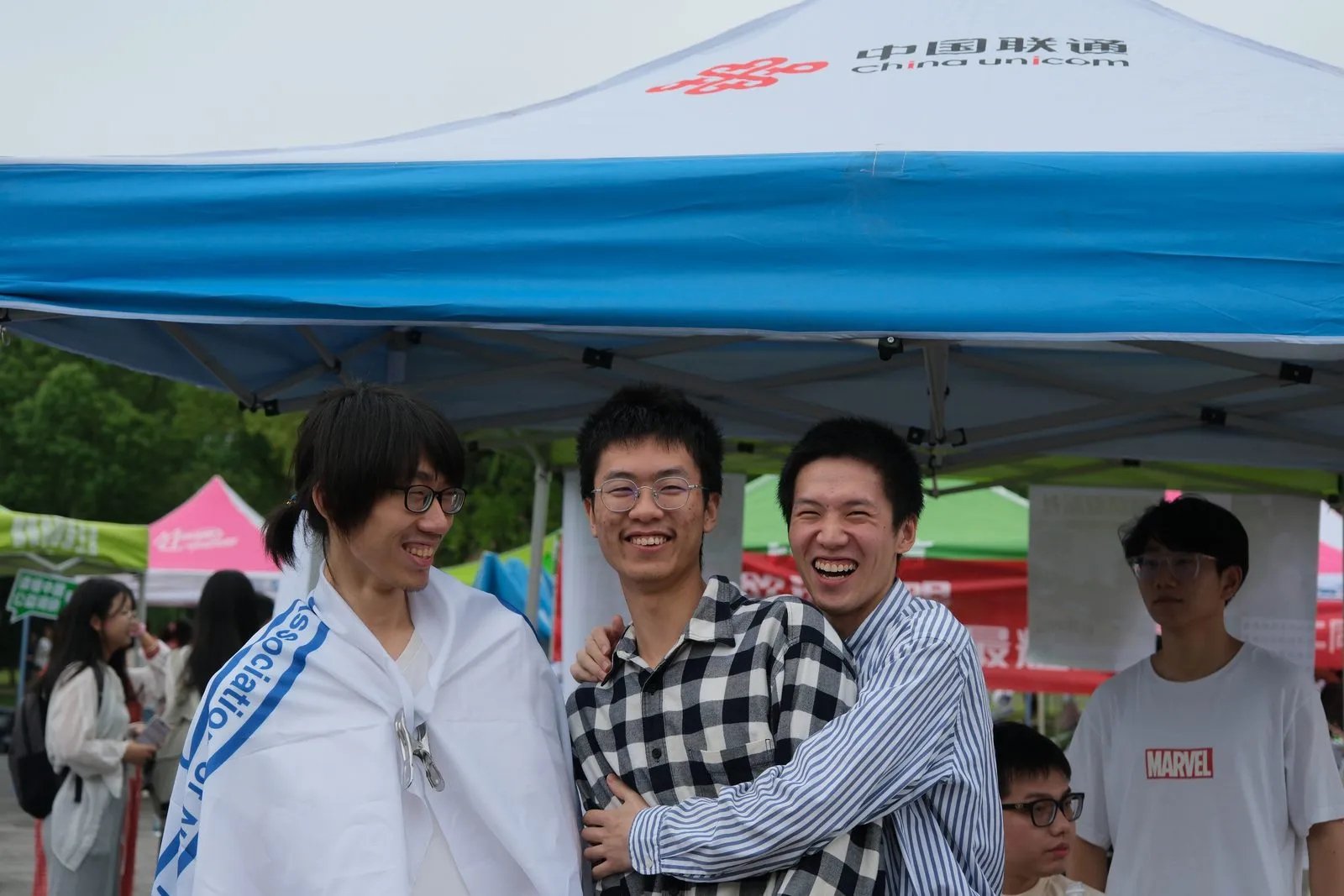百团大战现场，三名成员在蓝白帐篷下搂着肩大笑，其中一人披着印有 Association 字样的绶带，背景是其他社团的粉色与绿色摊位