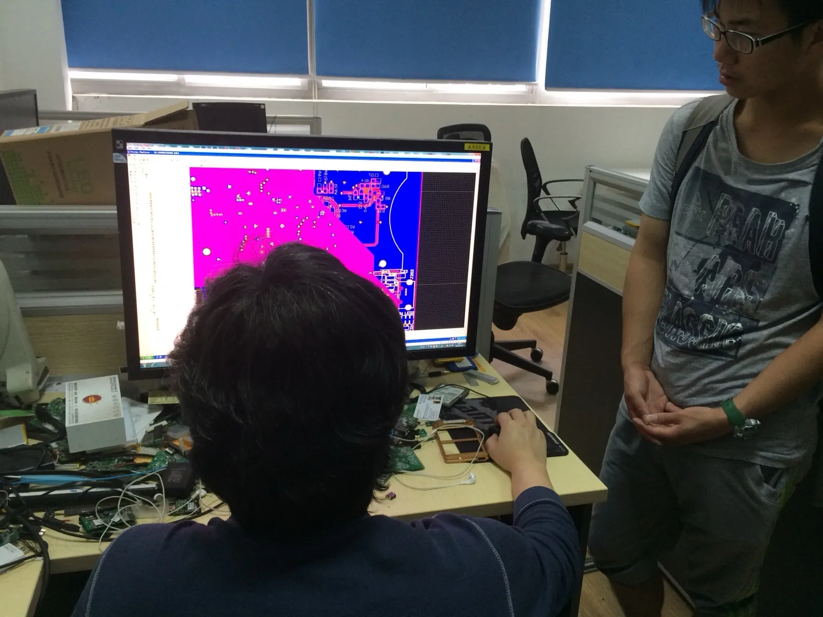 办公室工位上，一名工程师背对镜头坐着，面前的显示器上是 PCB 设计软件的布线视图，粉紫与蓝色的铜层叠在一起；桌面散放着多块拆下的电路板、螺丝刀与一块电池，右侧一名背着双肩包的学生站着看屏幕