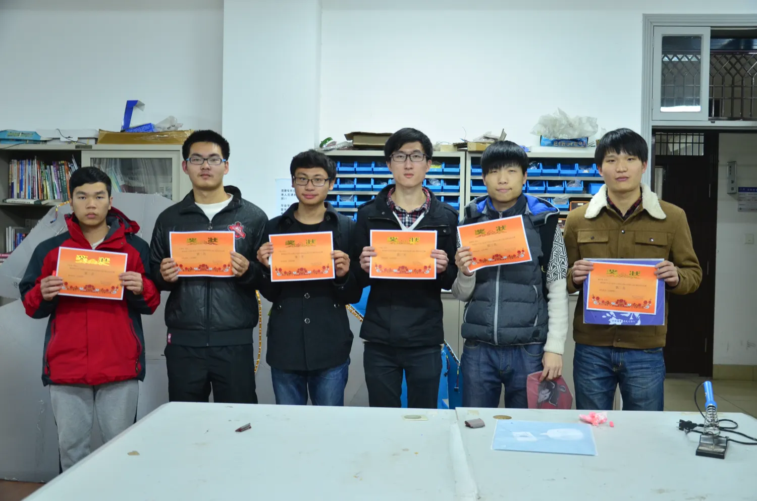 六名学生并排站着，每人双手举起一张橙色奖状，奖状上方印“奖状”二字，下方一行小字标着名次；背景是实验室的书架与一排排蓝色零件盒，桌上放着电烙铁与工具