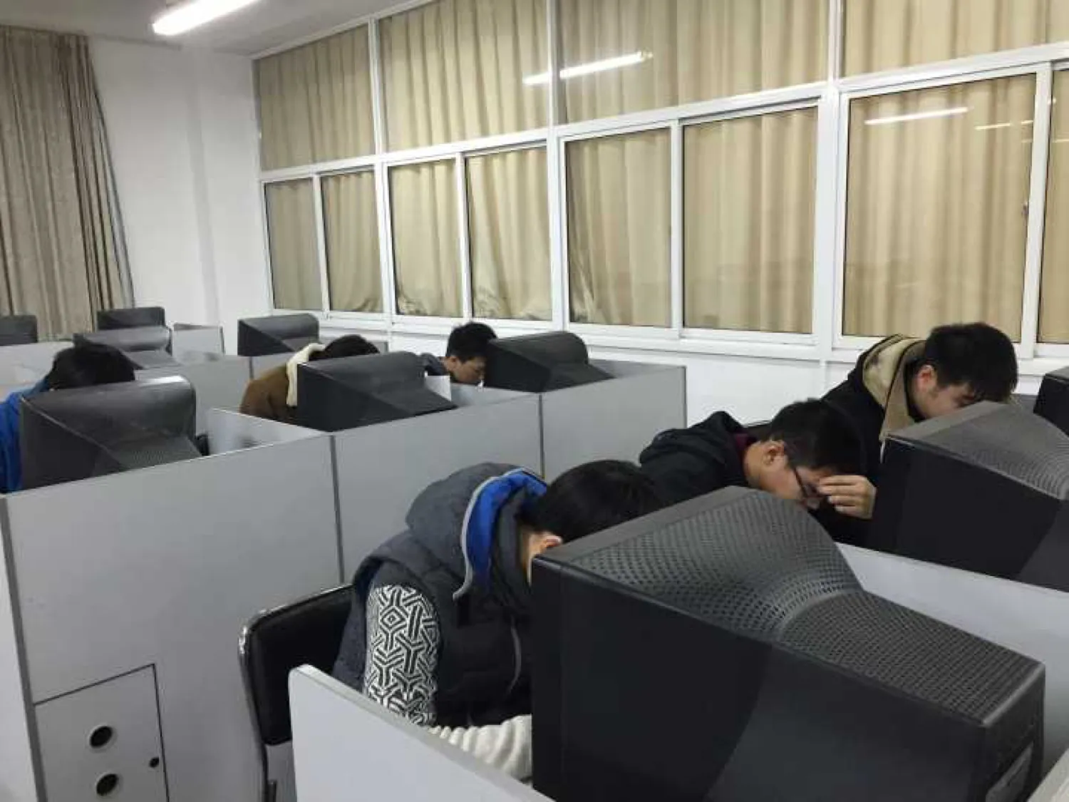 机房里一排排带隔板的机位，参赛学生分坐其间对着显示器答题，多数人穿着冬季外套，靠窗一侧拉着米色窗帘