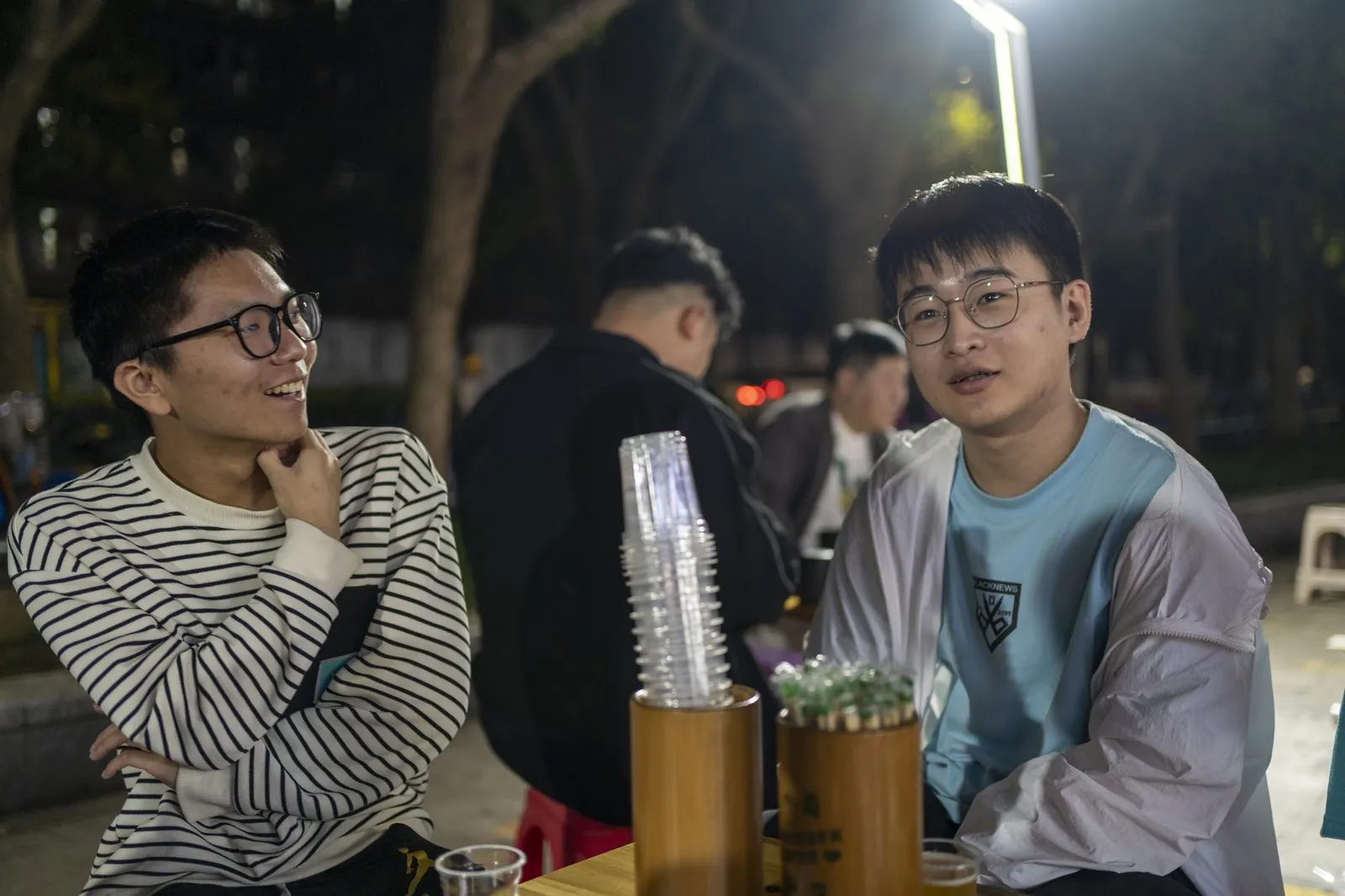 活动结束后的烧烤小聚，夜里户外长桌旁两名成员端着饮料聊天，背景是其他成员