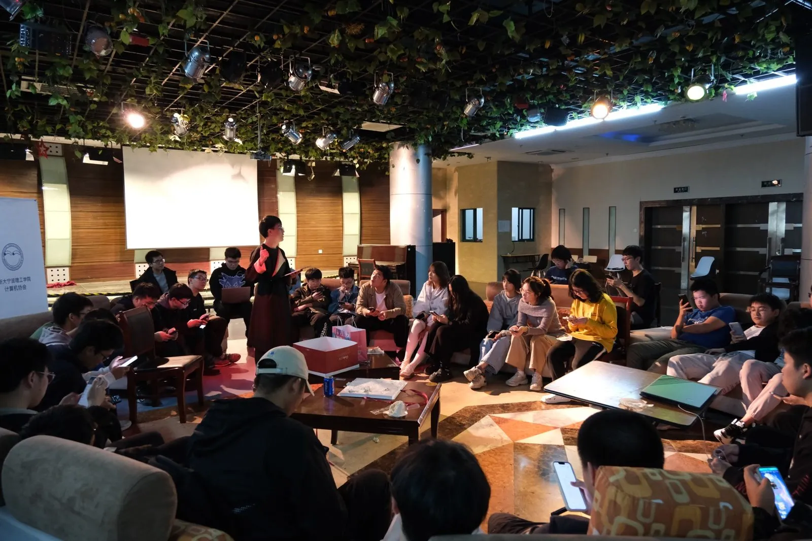 2023 年秋季迎新大会，成员围坐在沙发与茶几旁听一位成员讲话，左侧立着写有“浙大宁波理工学院计算机协会”的展架