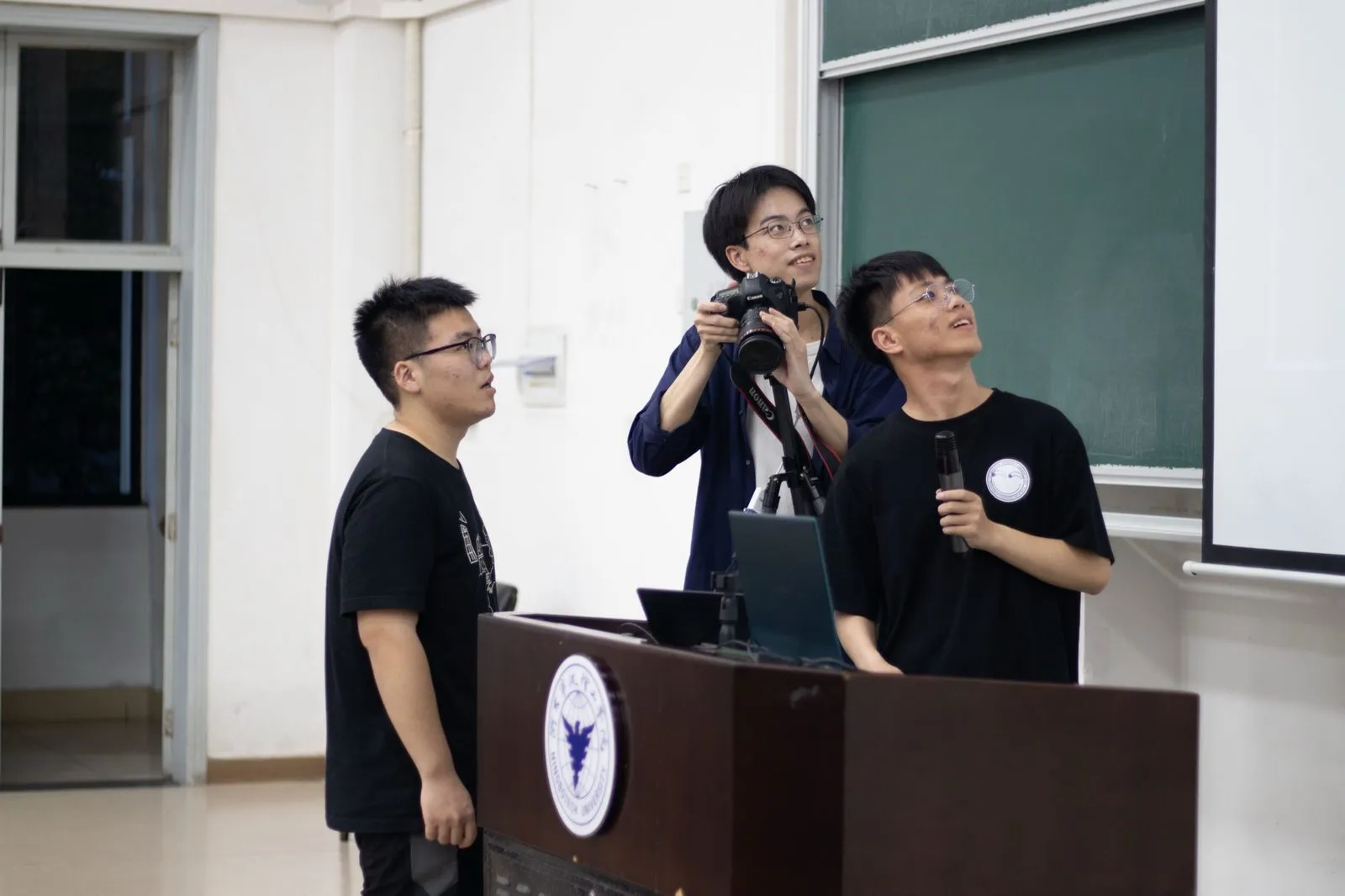 讲座现场，一名成员手持话筒在讲台前讲解，另一名成员架着相机拍摄