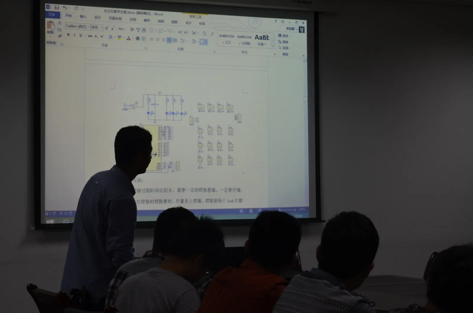 昏暗的教室里，投影幕上是一份名为“光立方教学方案.docx”的 Word 文档，页面上方为电路原理图，下方可见“焊接过程时间比较长，需要一定的焊接基础，一定要仔细”等文字；一名讲者背对镜头站在幕布前，前排几位学生仰头看着