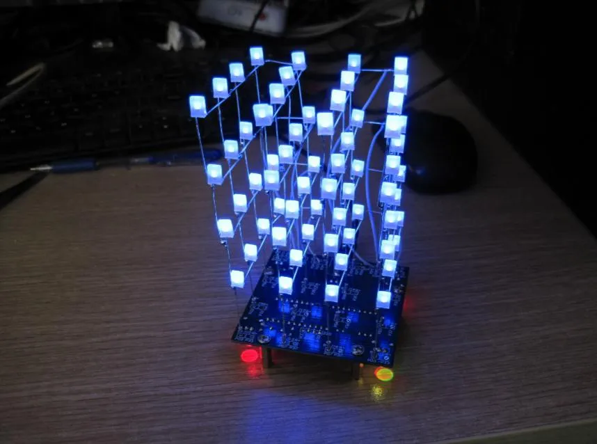 桌面上点亮的光立方：蓝色底板立起四层共 64 颗方形 LED，全部发蓝光，细导线在灯珠之间连成网格，底板边缘另有一红一黄两颗指示灯亮着，背景是键盘与鼠标