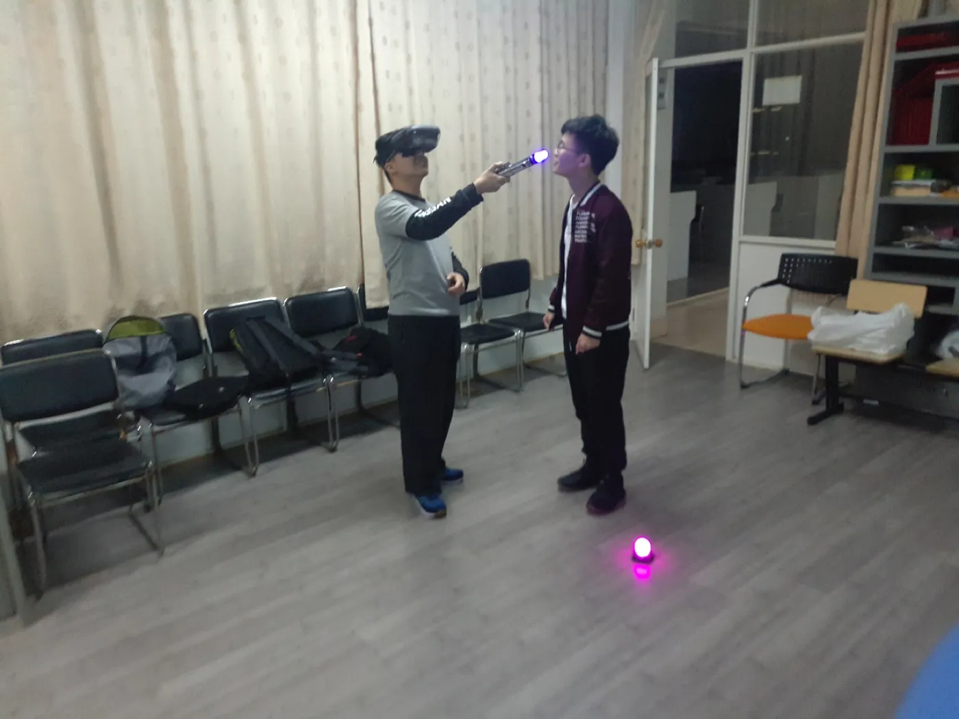 室内空场地上两名学生相对而立，一人戴着头显举起发光的光剑指向对方，另一人侧身看着；地面上有一枚发出紫光的圆形追踪信标，背景是一排排会议椅