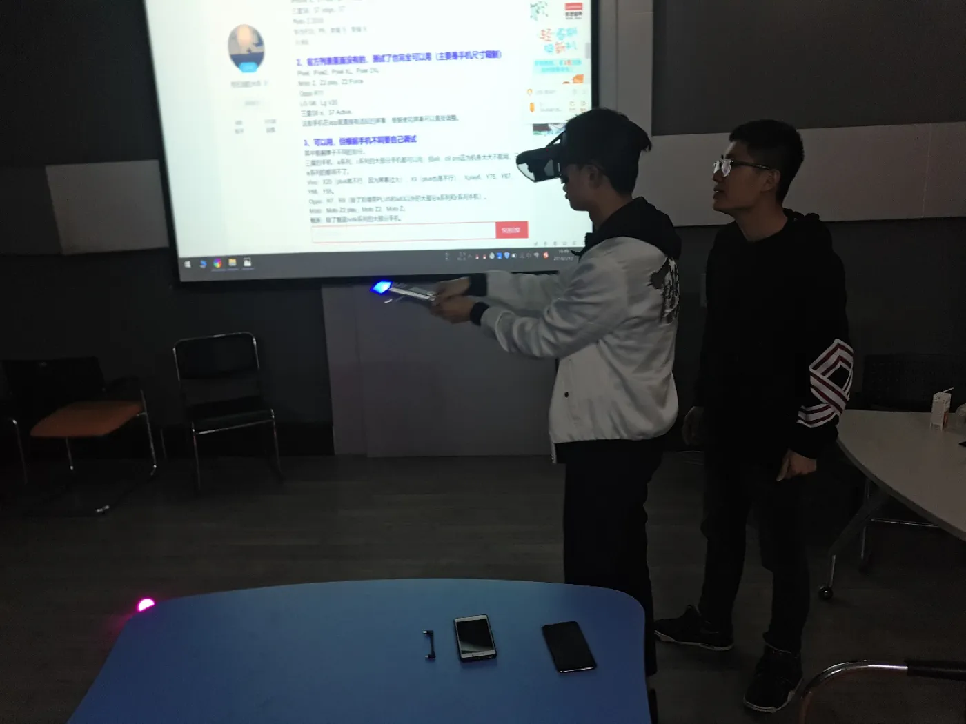 一名学生戴着 Mirage 头显、手持发亮的光剑站立，旁边一人陪着；两人身后的大屏幕上是一个网页，地面有紫色追踪信标，桌上放着两部手机