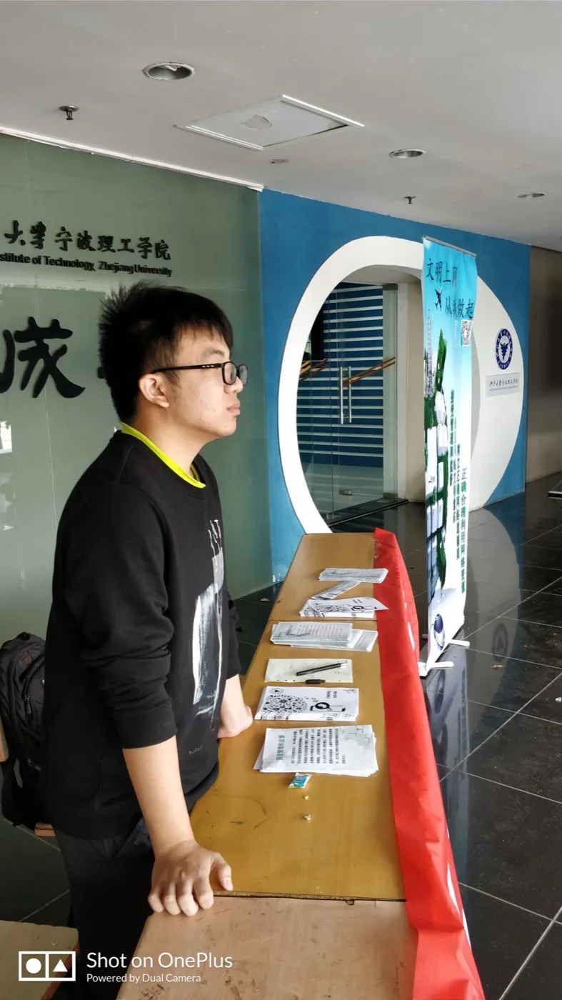 教学楼一楼大厅里,一名戴眼镜的学生站在铺着红布的长条桌旁,桌上分几摞放着宣传单,其中几张印有二维码;身后是一块蓝色易拉宝,上面写着“文明上网 从我做起”与“正确合理利用网络资源”,再往左是刻着“浙江大学宁波理工学院”的玻璃墙
