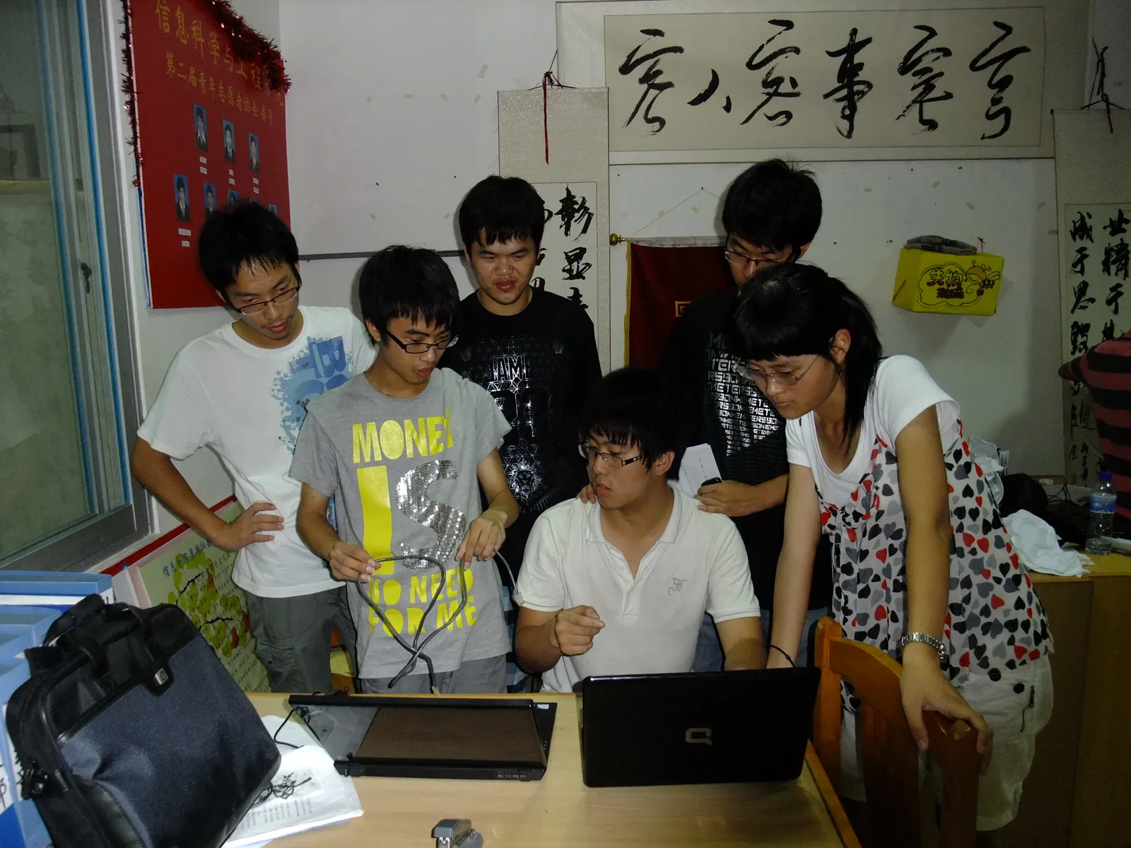 办公室里，一名成员坐在桌前操作笔记本电脑，另外五人站着俯身围看，其中一位手里拎着一根数据线；桌上还摊着一台合上的笔记本与一叠纸。墙上挂着一块贴有成员证件照的红色展板和几幅书法条幅