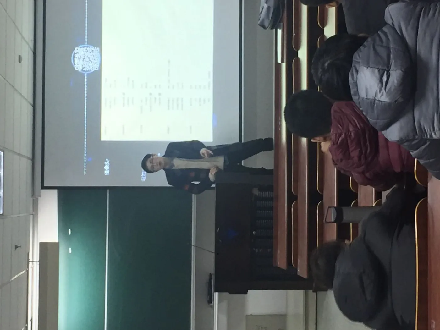 从教室后排隔着几名穿冬装的同学望向讲台，投影幕上是一页浅色幻灯片，一名讲者站在幕布前的讲台边讲解