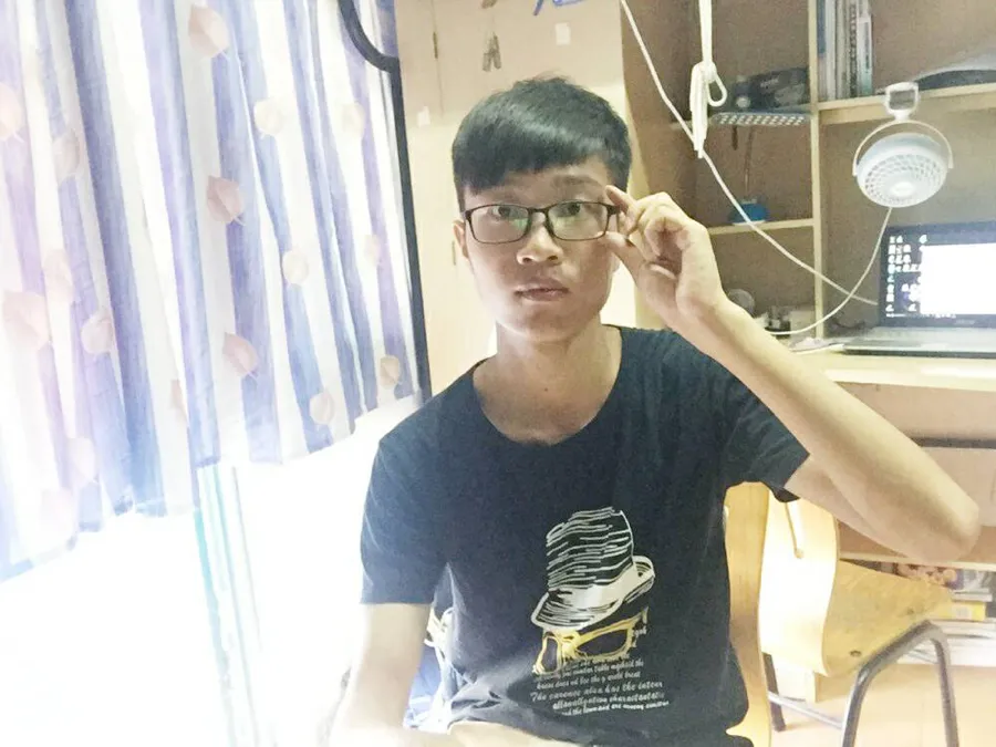 宿舍里的自拍：一名戴眼镜、穿黑色印花 T 恤的男生坐在书桌前，一手扶着眼镜，身后是台灯与书架