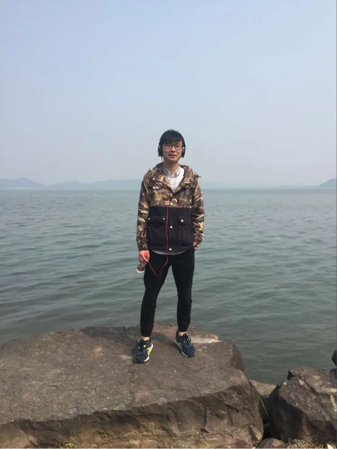 湖边的全身照：一名穿花衬衫的人站在一块伸入水面的岩石上，背景是开阔水面与远岸