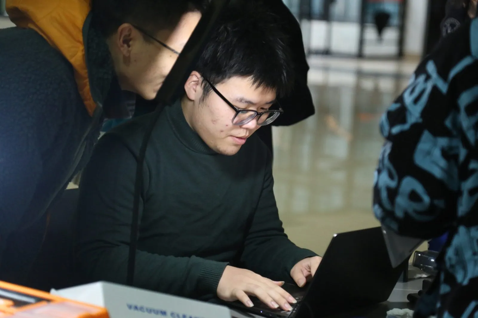 一名队员俯身在笔记本上操作，另一名队员在旁看着屏幕，桌上放着吸尘器包装盒