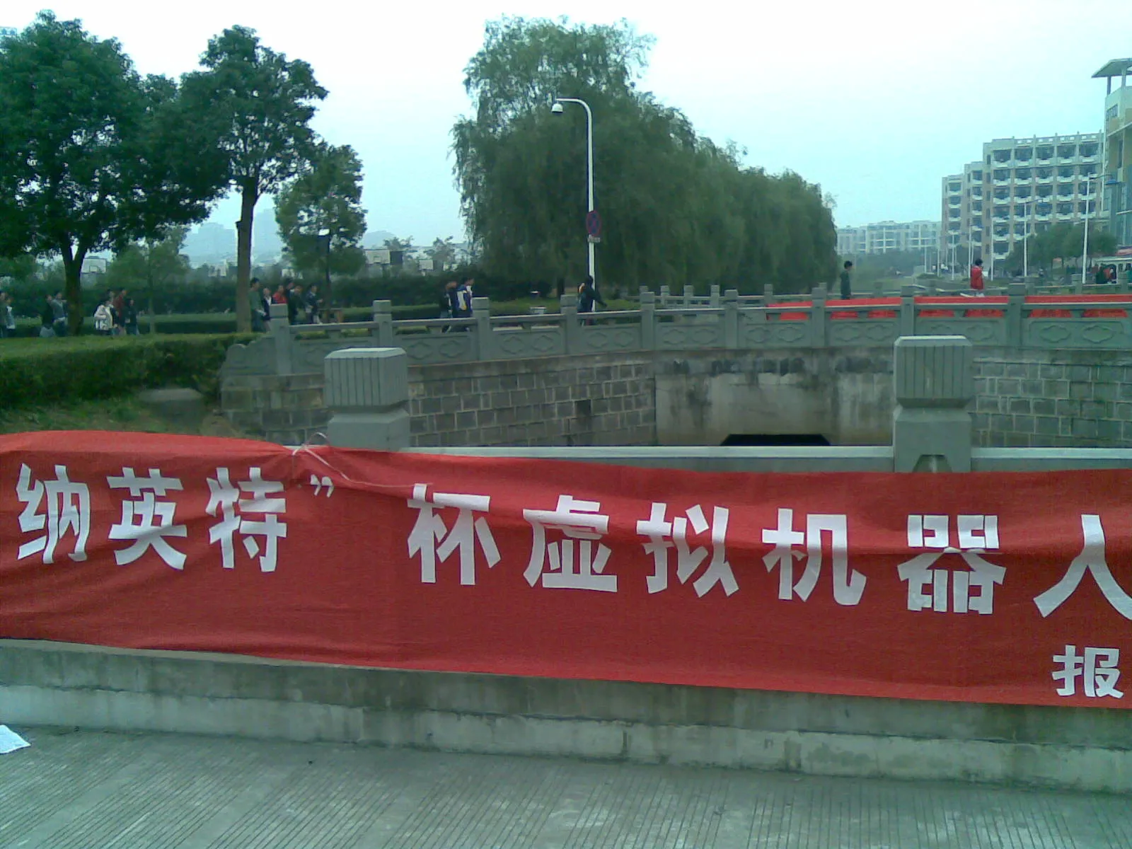 校园河道边的石栏杆下挂着一条红色横幅,白字写着“纳英特”杯虚拟机器人…报…,远处栏杆上还系着另一条红色横幅;背景是柳树、路灯与教学楼