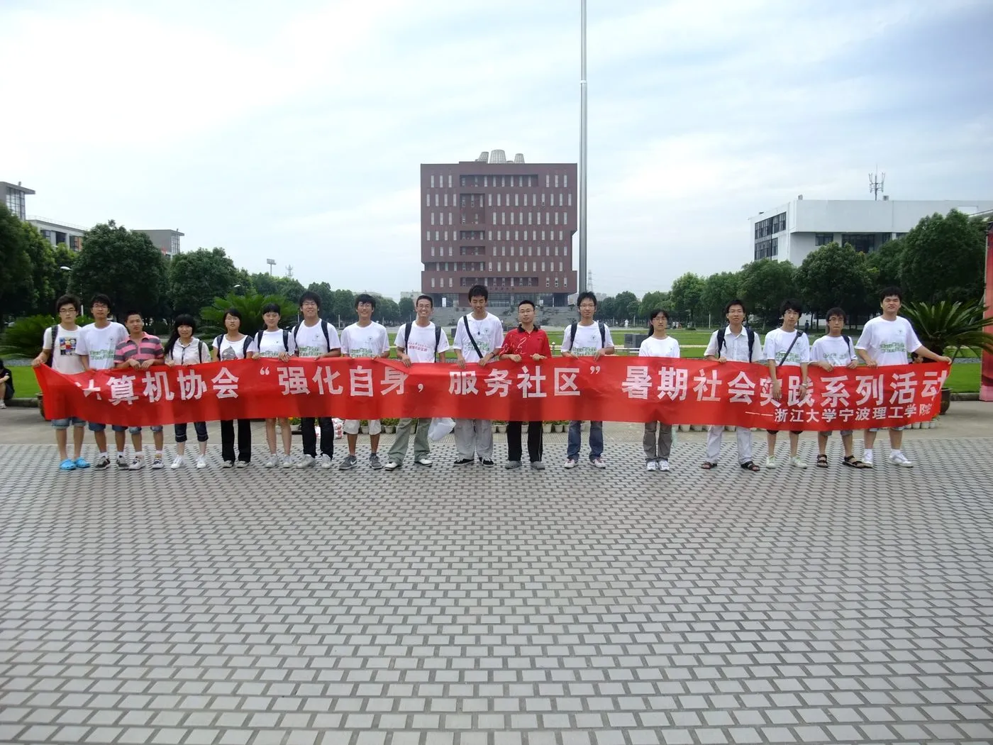 近二十名成员在校园广场上一字排开，共同拉着一条红色横幅，多数人穿着印字的白色队服；身后是旗杆、草坪与一栋红砖高楼。横幅写有“计算机协会‘强化自身，服务社区’暑期社会实践系列活动——浙江大学宁波理工学院”