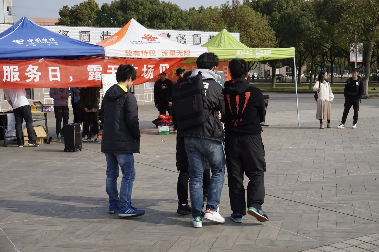 露天广场上支起三顶帐篷，分别印着中国电信、中国联通与一家运营商的 4G 广告；帐篷前拉着一条红色横幅，可见“服务日，露天广场”字样。几名学生背对镜头站在场地中间，远处有人在摊位后整理物品