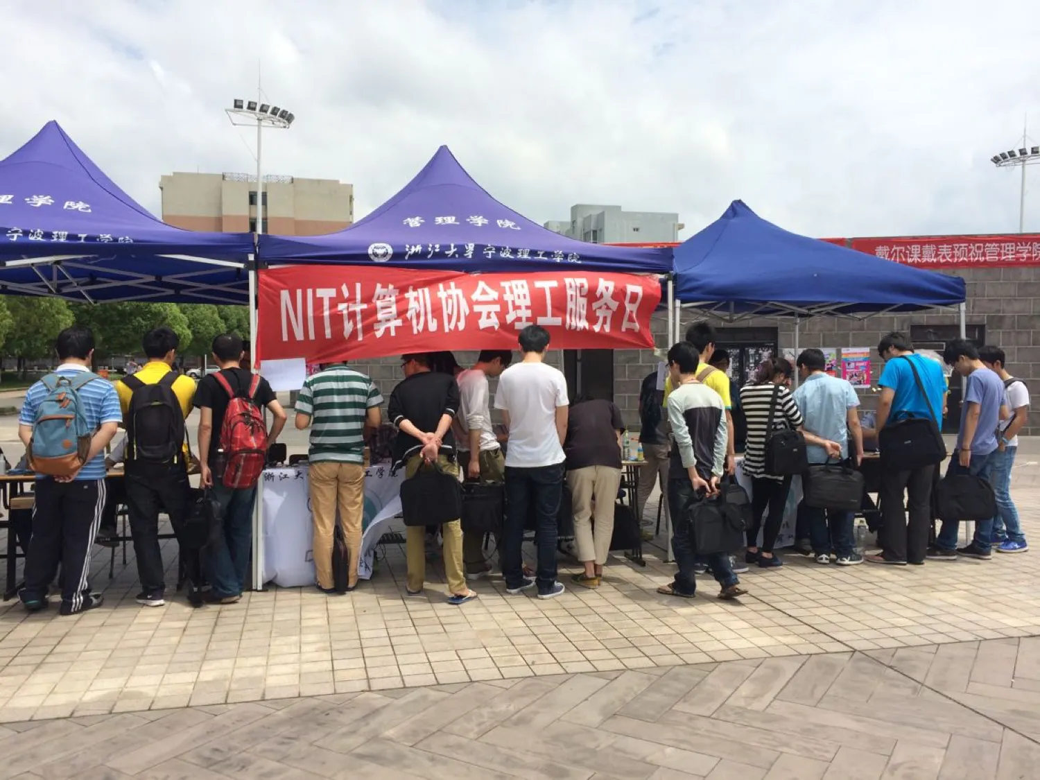 露天广场上并排支着几顶蓝色帐篷，帐篷顶印着“浙江大学宁波理工学院”与“管理学院”；前方拉一条红色横幅，写着“NIT计算机协会理工服务日”。二十来名学生背着包排在长桌前等候，右侧另一顶帐篷的横幅上可见“戴尔课戴表”字样