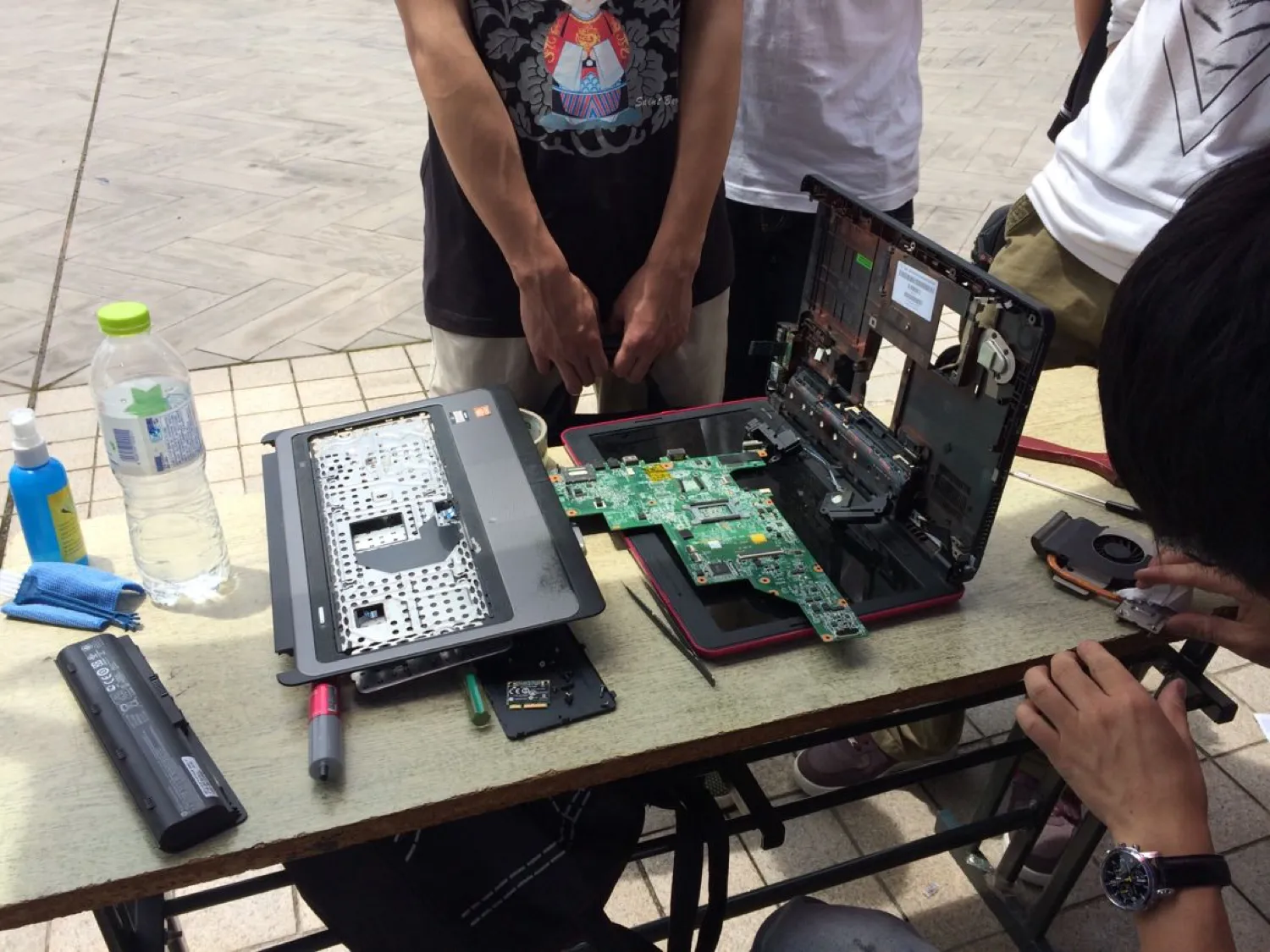 长桌上摊着一台拆到底的笔记本：上盖、卸掉键帽的掌托、单独取出的主板、电池、风扇分开摆着，旁边放着撬棒、清洁喷雾与一块蓝色擦布；两名同学站在桌前看，右侧一只手正在操作