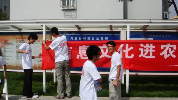 五名穿白色队服的学生在一排村务公开栏前挂一条红色横幅，横幅上“息文化进农”几个大字露在外面；公开栏的蓝色标题写着“湾底村村民自治章程”，背景是村里的楼房
