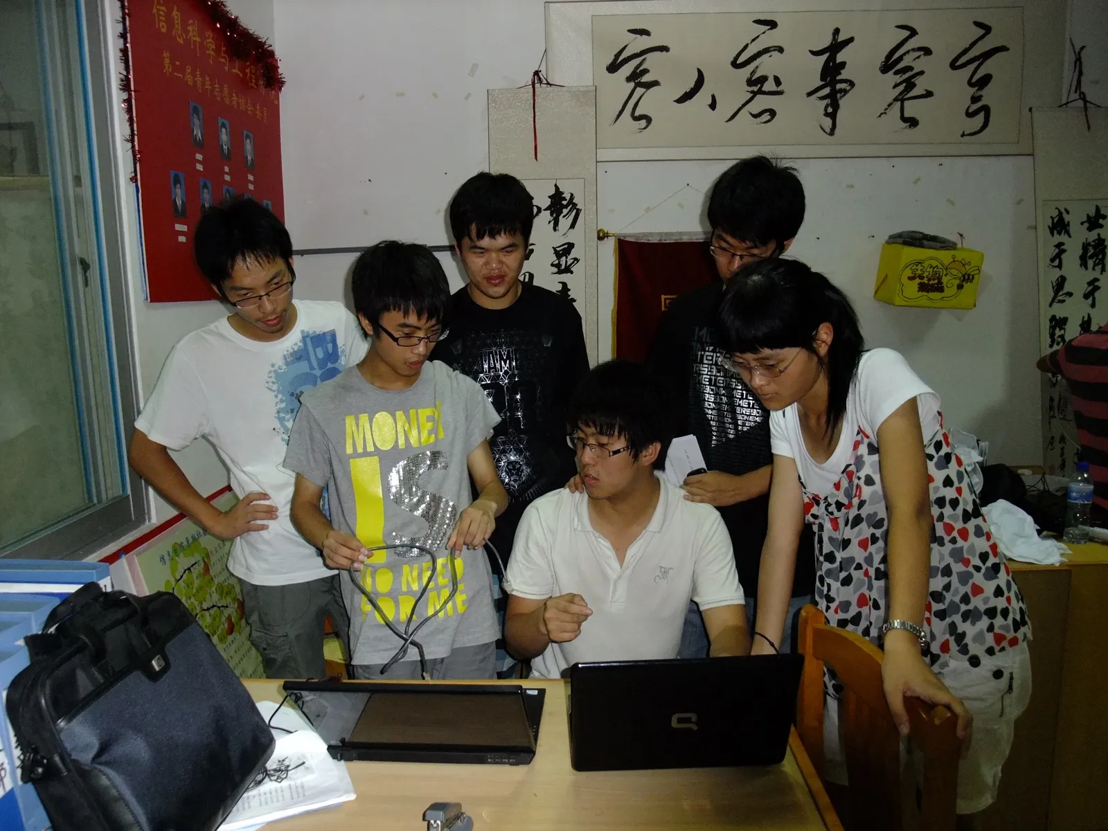 六名学生挤在一张办公桌前，围着两台笔记本电脑。坐着的一位在操作，旁边一位手里拎着一根数据线正在解释，其余人俯身看屏幕。墙上挂着“信息科学与工程学院第二届青年志愿者协会”的红色展板与几幅书法