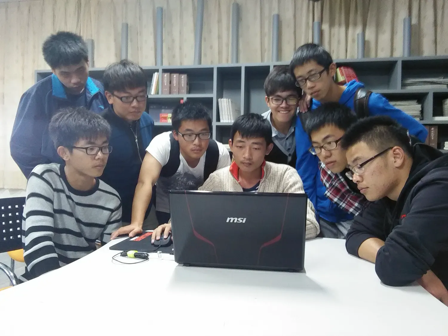 八九名学生紧紧围成一圈，挤在一台 MSI 笔记本前；居中一人手放在触控板上操作，其余人俯身盯着屏幕，背景是书架
