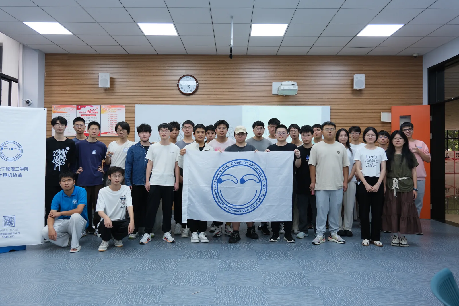 室内合影：二十余人站成两排，多数比出手势或笑着看镜头；左侧立着一块白色易拉宝，上面是协会的圆形笑脸标识