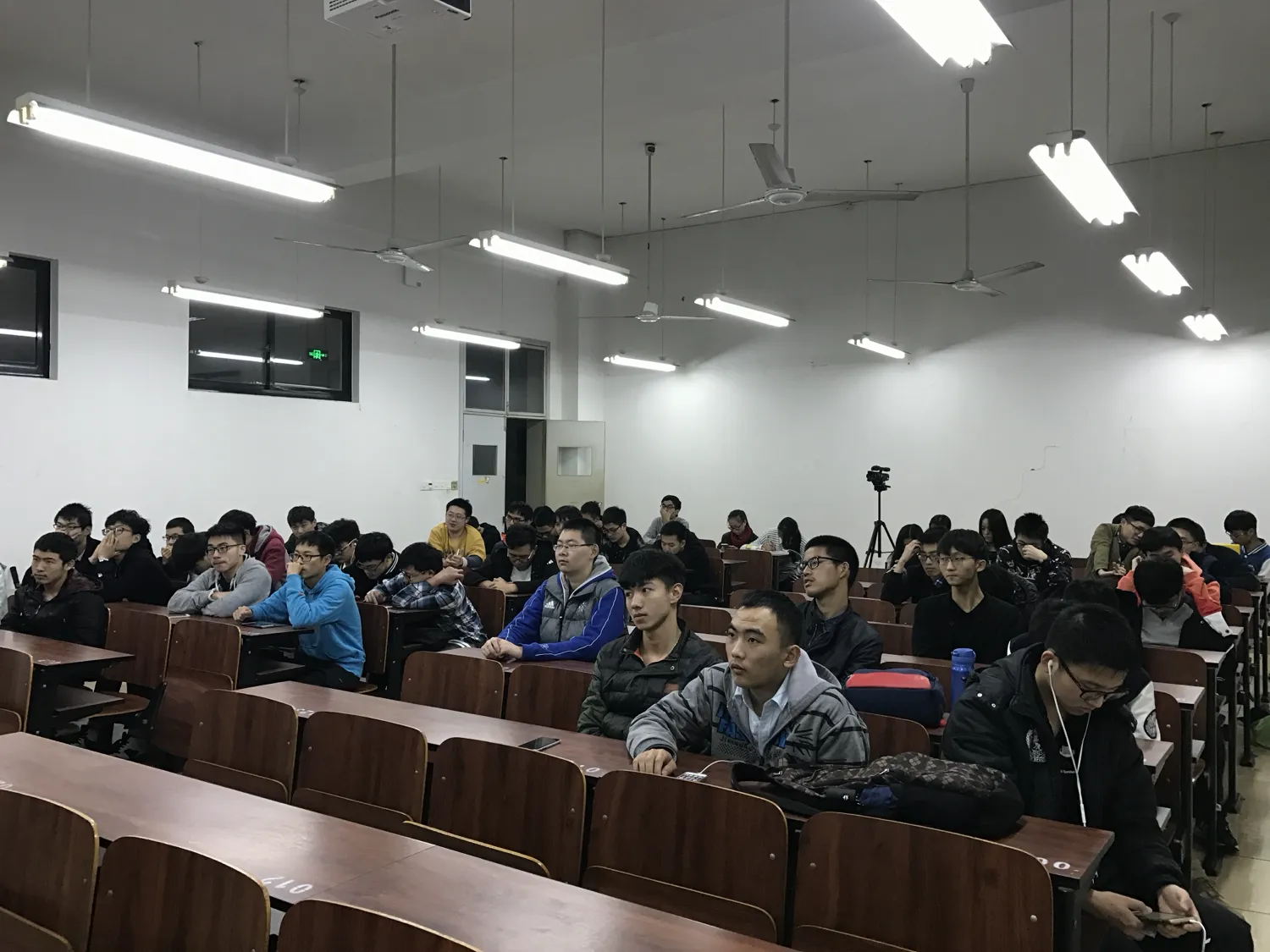 阶梯教室里坐满穿冬装的学生，一直坐到最后一排；靠右过道处架着一台三脚架上的相机，前方讲台亮着