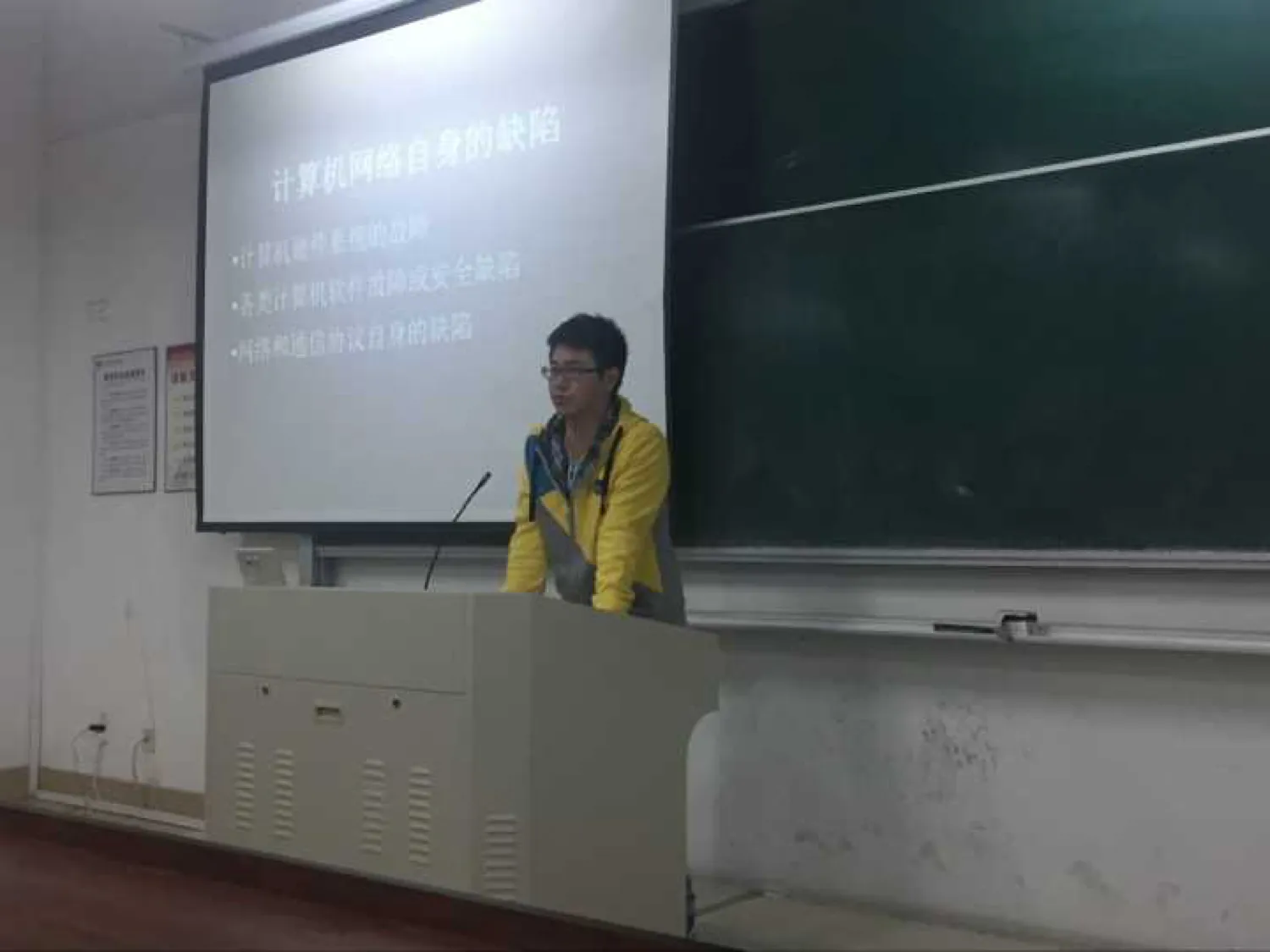讲台前一名穿黄色外套、戴眼镜的讲者站在话筒后；身后投影幕上是一页标题为“计算机网络自身的缺陷”的幻灯片，下列三条：计算机硬件系统的故障、各类计算机软件故障或安全缺陷、网络和通信协议自身的缺陷